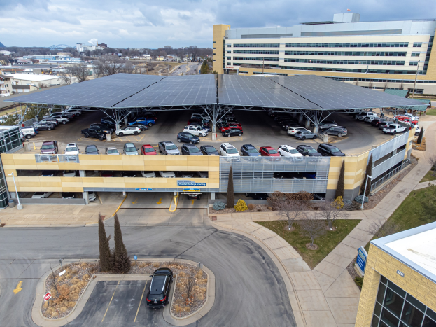 Luftaufnahme eines solarpanelbedeckten Parkplatzes mit parkenden Autos, umgeben von Gebäuden, Bäumen, Grüss, Straße und Höhen unter einem bewölktem Himmel.
