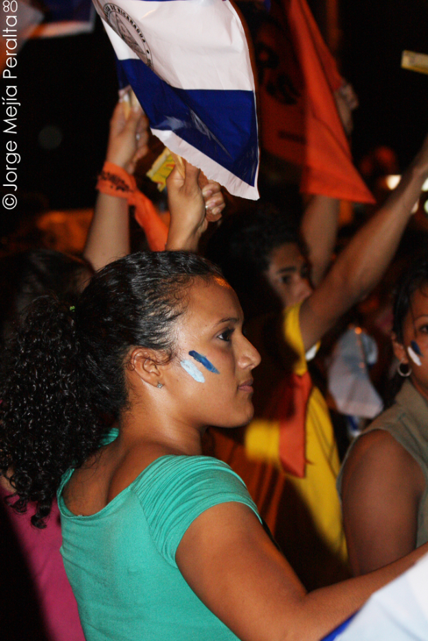Eine Frau in einem gr√ľnen T-Shirt steht auf einer Demonstration und h√∂lt ein weißes Schild, umgeben von vielen anderen Menschen.