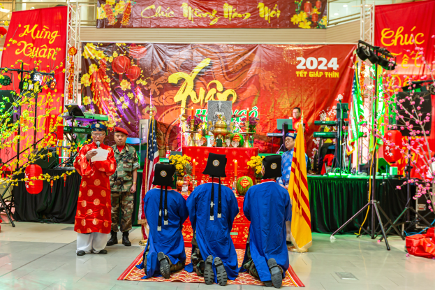 Eine Gruppe von Menschen sitzt auf einem roten Teppich vor einer Bühne mit Bannern, Lichtern und Blumen, während eine Person ein Papier hält, wahrscheinlich bei einer Chinesischen Neujahrsfeier in Singapur.