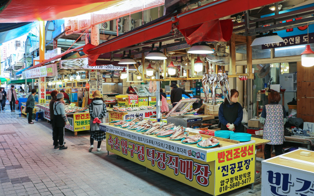 Ein belebter Straßenmarkt in Seoul, Südkorea mit Menschen, die gehen, Tischen mit verschiedenen Gegenständen und Gebäuden unter einem klaren blauen Himmel.