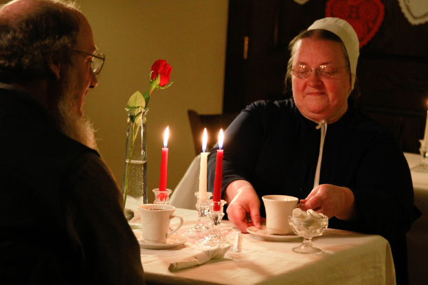 Zwei Personen sitzen an einem Tisch mit einem Becher, Kerzen und einem Blumenarrangement.