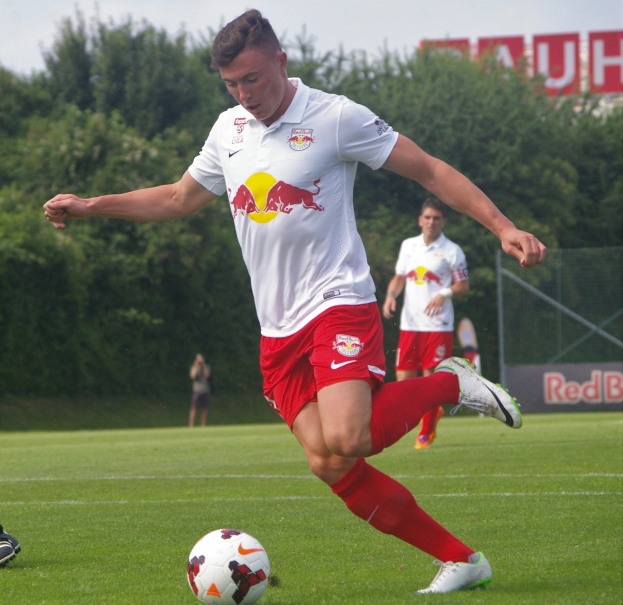 Ein Mann in einem Fußballtrikot schießt einen Ball auf einem grünen Feld mit Bäumen und einem klaren blauen Himmel im Hintergrund, mit einem Schild mit der Aufschrift "RB Leipzig".