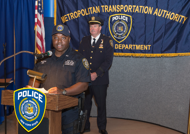Zwei Polizeibeamte stehen vor einem Rednerpult, einer hält Papiere und spricht in ein Mikrofon, mit einer Tafel, einer Fahne und einem Tisch in der Nähe, und einer Tafel mit Symbolen und Text im Hintergrund.