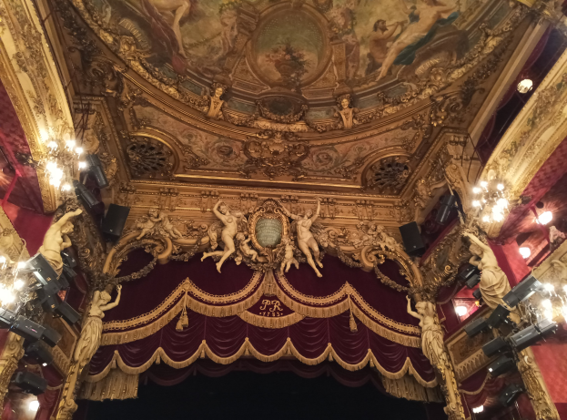 Theaterinnenraum mit roter Gardine, Kronleuchtern, Skulpturen, Lichtern und einer bemalten Decke, mit einer Bühne mit ein paar Menschen unten.