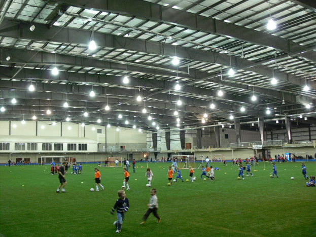 Eine Halle mit einer Gruppe von Menschen, die mit einem Fußball auf dem Boden spielen, Torpfosten und Beleuchtung auf dem Dach.