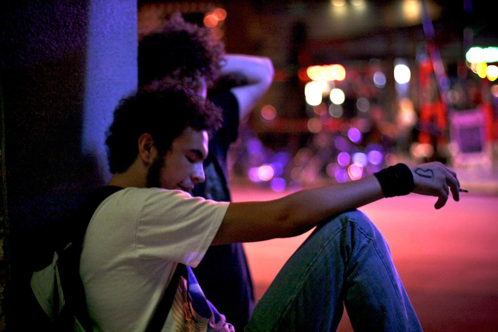 Mehrere Personen sitzen auf dem Boden, wobei eine Person eine Zigarette und eine Tasche in den Händen hält.